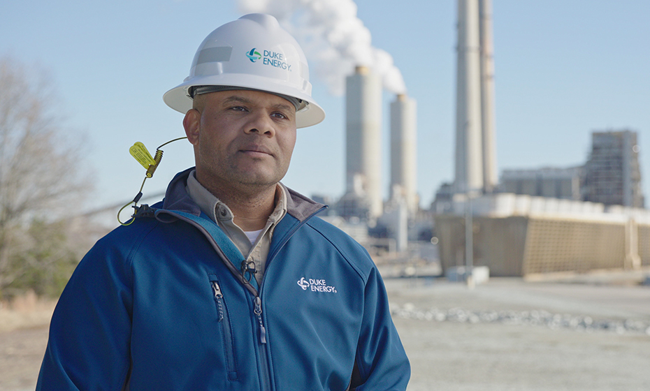 Derrick Smith, a chemistry technician at Duke Energy's Roxboro Steam Station, is pictured in front the of the plant. Smith wears a hard hat and a blue jacket.