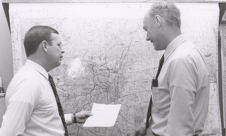 Two men stand in front of map while looking at a piece of paper.