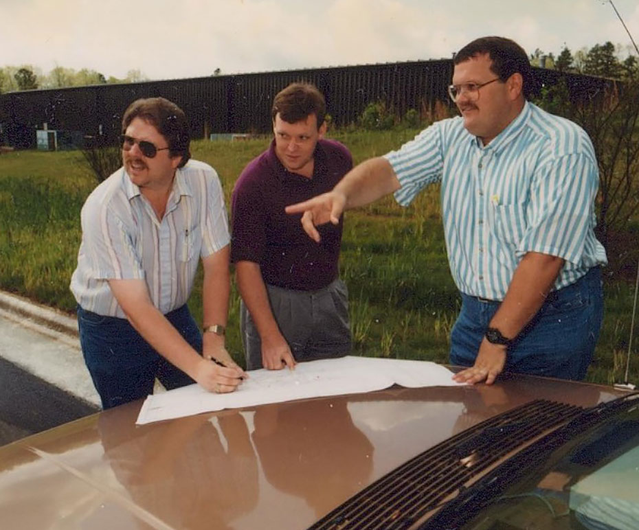 Three men stand in front of a vehicle. One man points to something off camera.