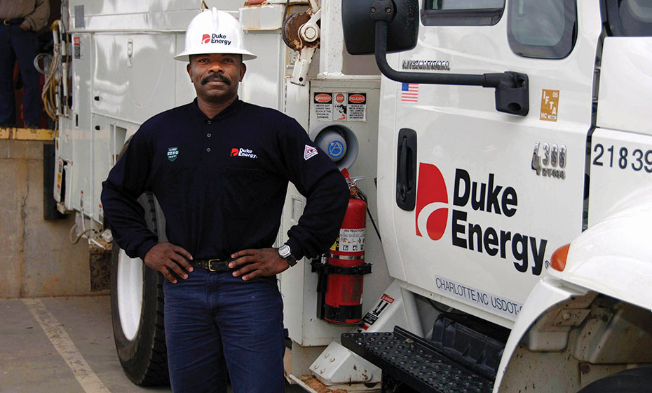 A man in a hard hat stands in front of a bucket truck as he smiles for a photo.