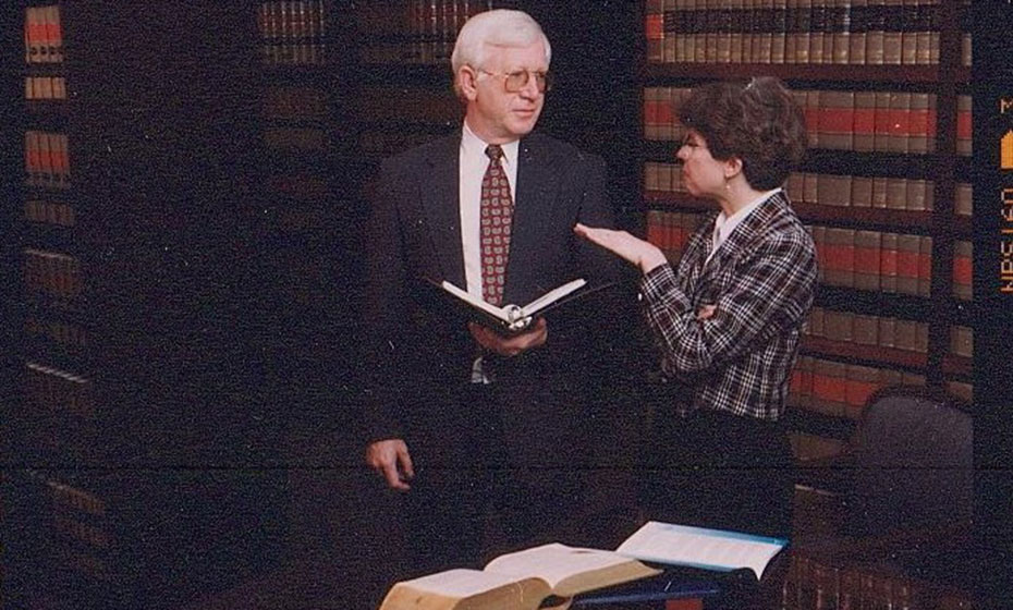 A man and a woman stand in a library as they engage in a discussion.