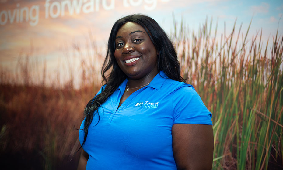 A confident Black female smiles for a photo, wearing a blue Duke Energy t-shirt that contrasts her long, dark hair.