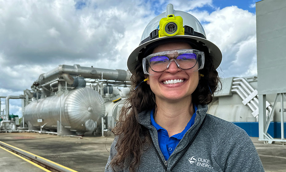 Taylor Bland wears a white hard hat and safety glasses as she stands in front of a Duke Energy power plant. She has curly brown hair and a broad smile.