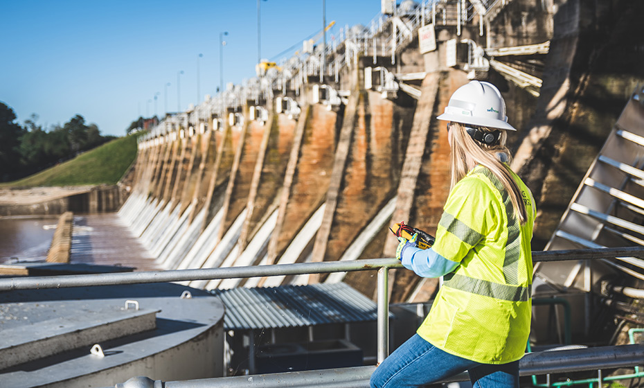 A female Duke Energy employee with blonde hair and a yellow safety vest stands in front of Tillery Hydroelectric Station in North Carolina.