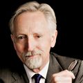 Headshot of Kim Burchiel, M.D., a man with grey hair and beard wearing a suit, holding his glasses in his hand near his face