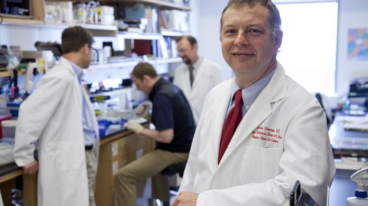 Researchers work in the background, while Markus Grompe, M.D. - a white man in a lab coat - stands in the foreground.
