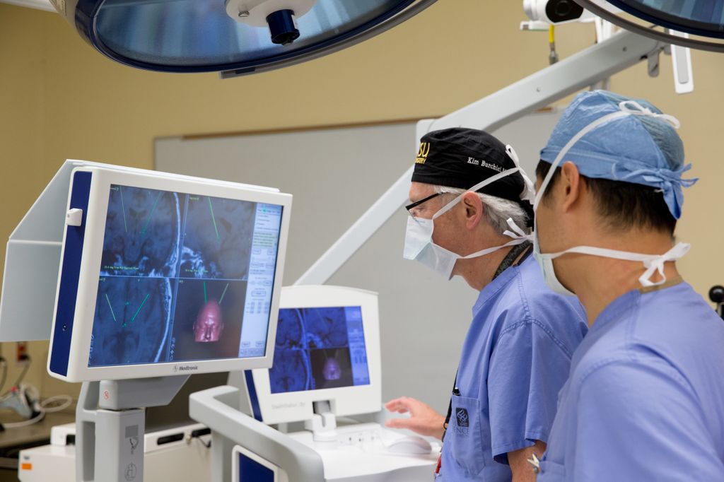 Two male doctors in scrubs view colorful displays on two computer monitors during deep brain stimulation