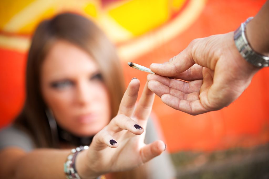 close shot of a person's hand offering a marijuana cigarette, and a teen's hand reaching for it, with teen out of focus in the background