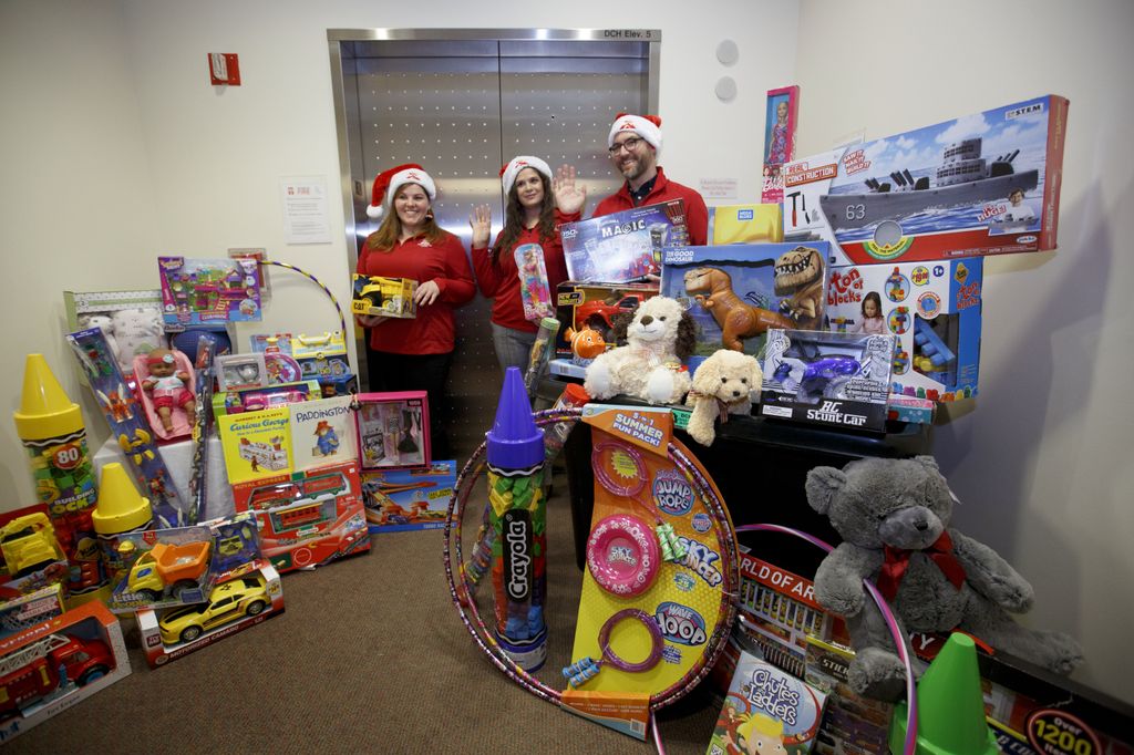 three people in santa hats waving from behind piles of toys