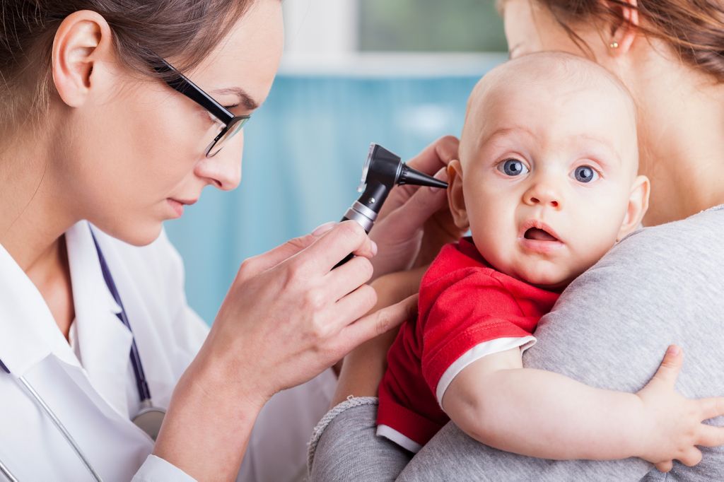 medical person looking in baby's ear, child resting on mother's shoulder looking at camera with a puzzled look