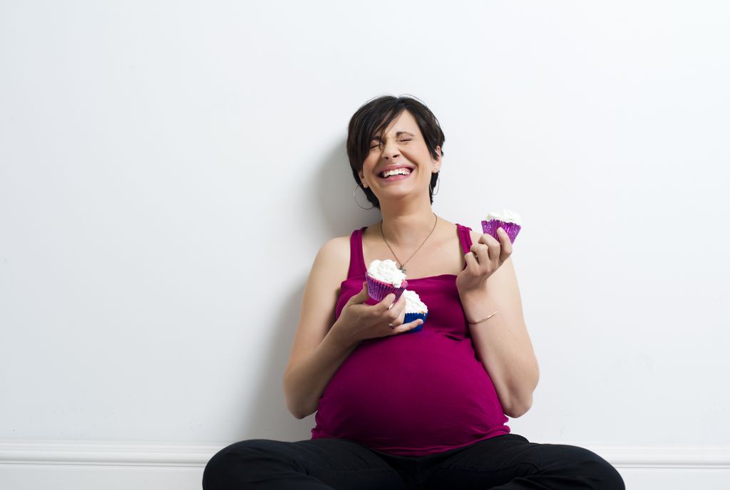pregnant woman seated on floor, laughing, holding 2 cupcakes