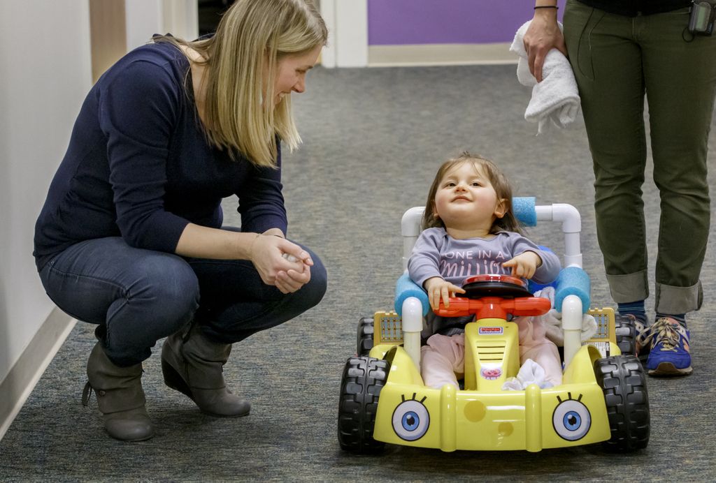 woman squatting next to a toy car, with a child inside the vehicle who is smiling