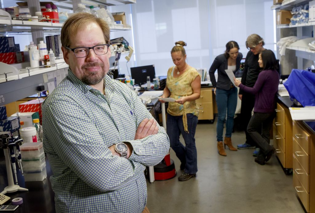 man in plaid shirt, arms crossed, smiling at camera while researchers work in background