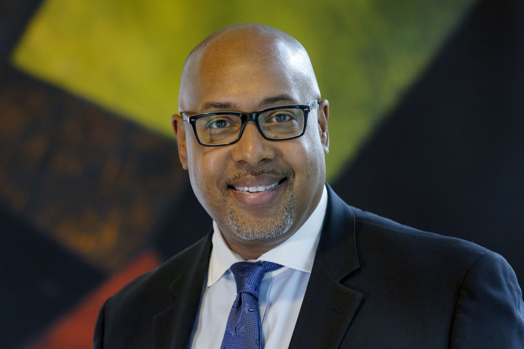 man with shaved head, glasses, in suit and tie, smiling at camera