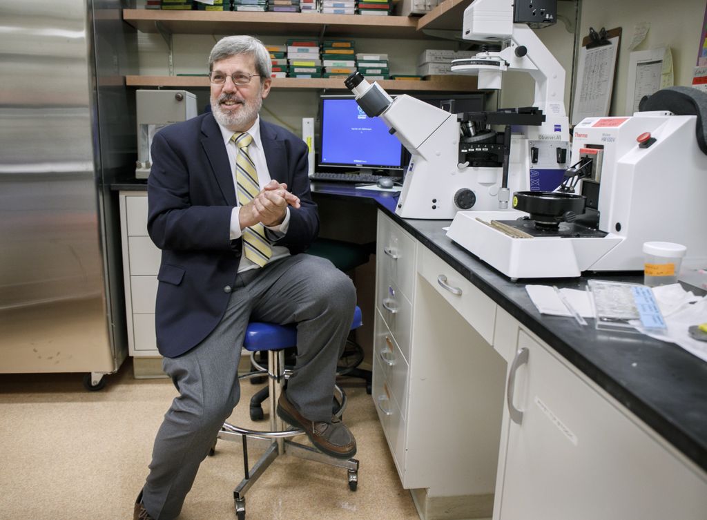 man in suit and tie, sitting in front of microscope, smiling 
