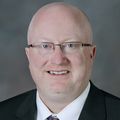 Joe Ness, M.H.A., BSPharm, is a person with no hair, glasses, and a suit, smiling.