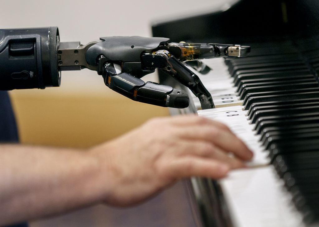 view from the side of a mechanical hand next to a person's hand, both playing the piano
