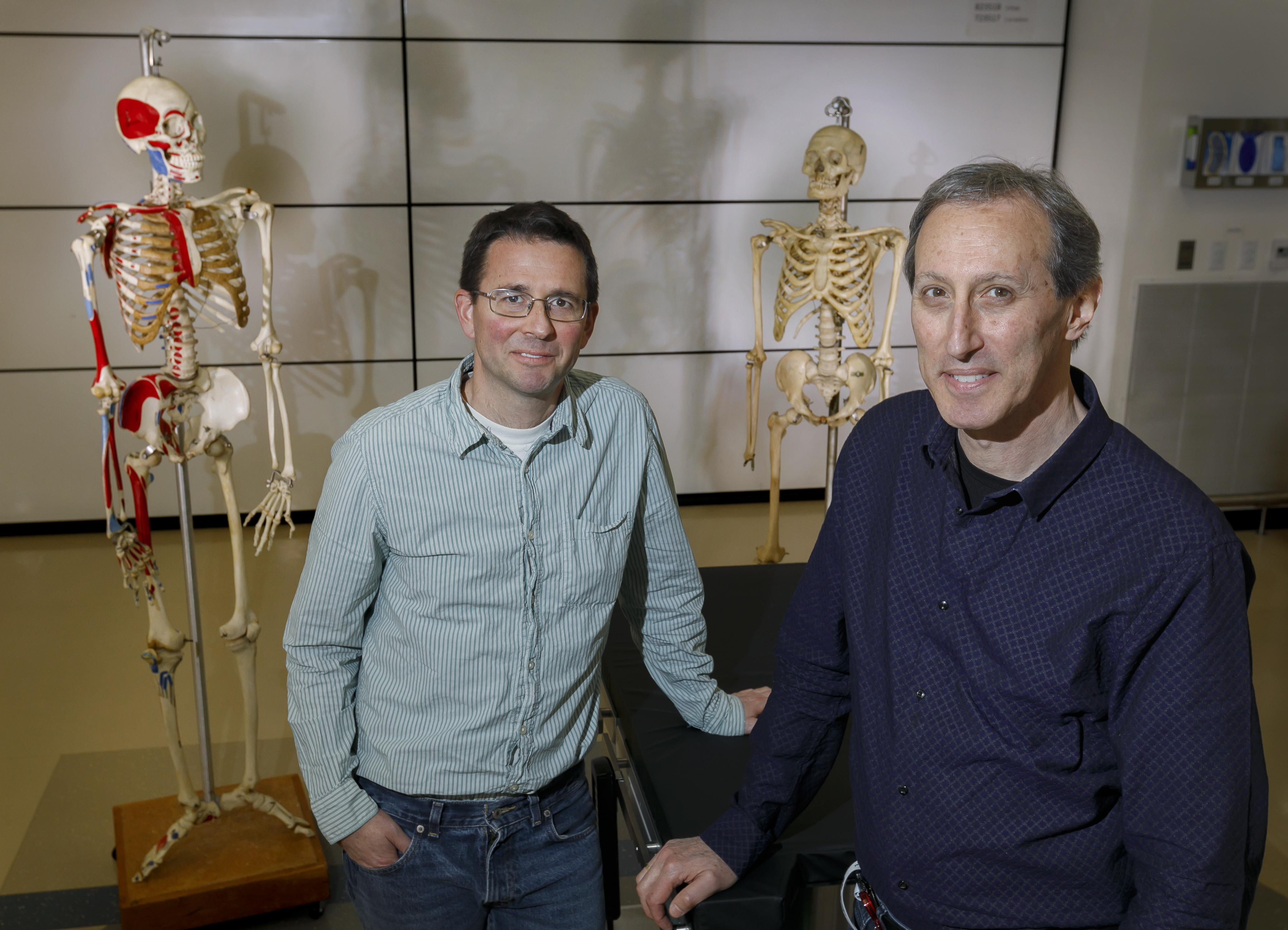 two men, standing around a medical examination table, with full-size skeletal models behind them