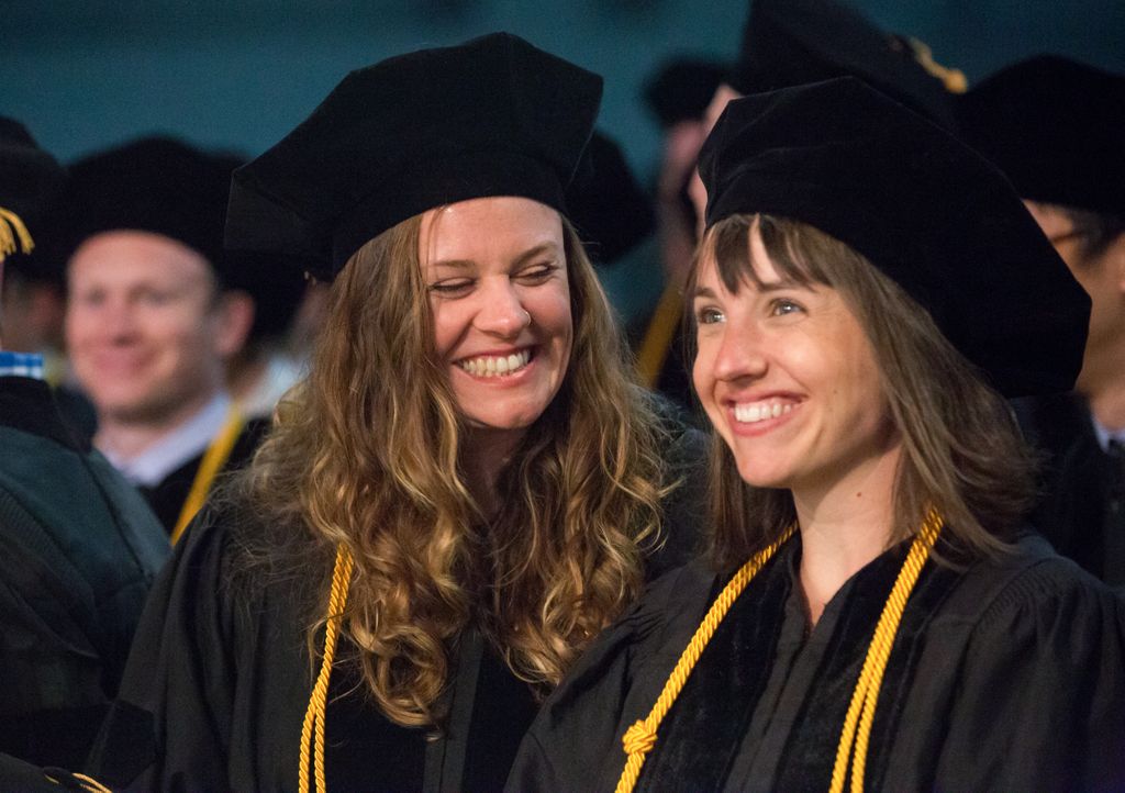 two women, smiling at one another, wearing graduation gowns and mortar boards