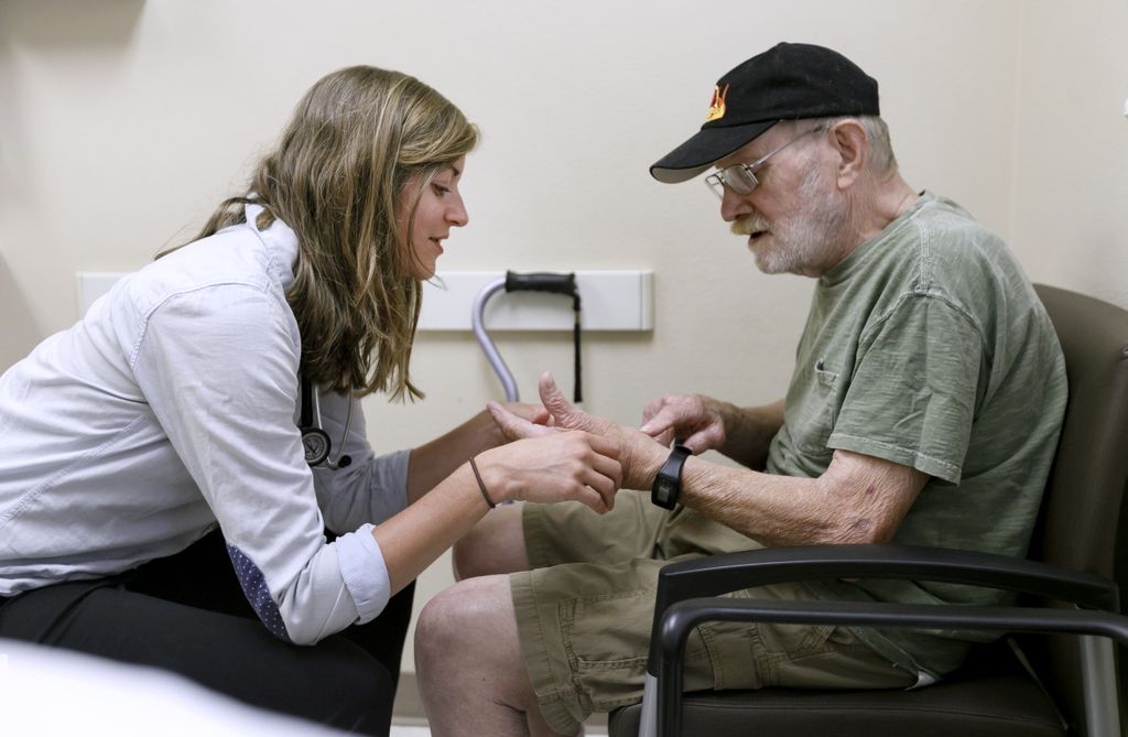 young female doctor, sitting face to face with a senior man as she looks at his hands
