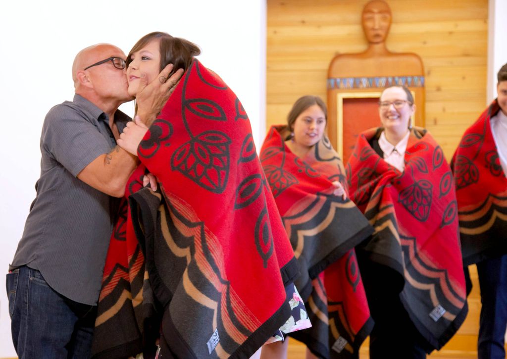 A young woman wears a red patterned wool blanket over her shoulders as a bald man kisses her on the cheek and two other young women also wearing red wool blankets watch behind them.