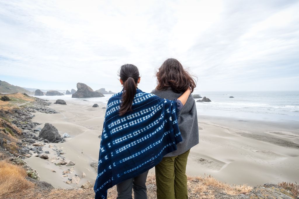 two women wrapped in blankets, standing with arms around one another on a beach, seen from behind