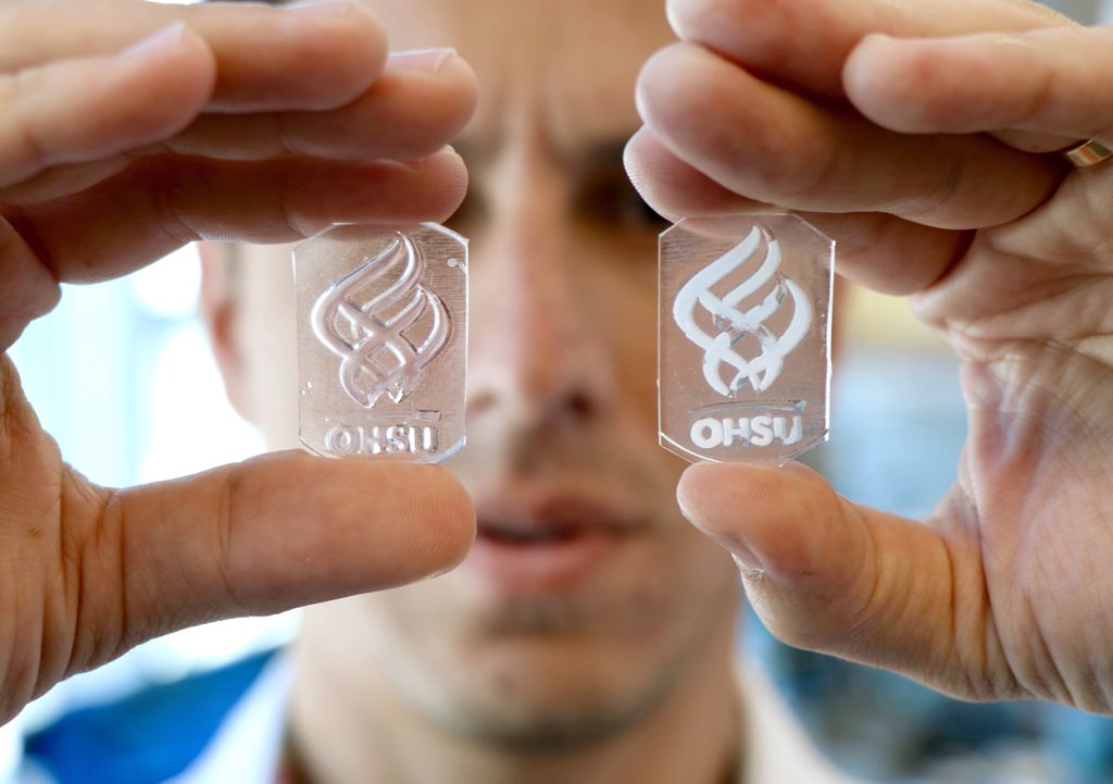 close up photo of bertassoni holding two slices of engineered material with the ohsu logo, one in each hand, in front of his face, with face out of focus in background