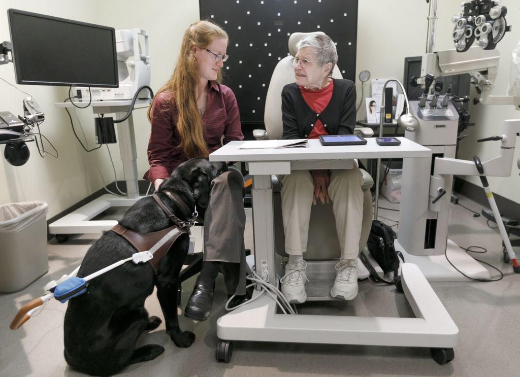 kathryn sits on left, helping beverly on right, teaching her to use a reader to read a newspaper, as kathryn's seeing eye dog nabisco sits at kathryn's fit on the left