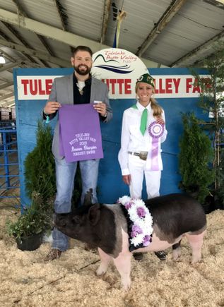 quiana, right, dressed in white and holding up a ribbon as she stands behind a large show pig at the fair