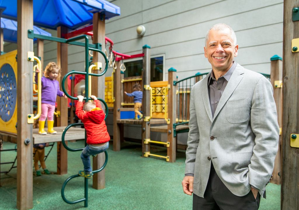 older gentleman with short gray hair wearing a gray suit with hands in pockets, smiling at camera, standing on playground with children playing on playground equipment behind him