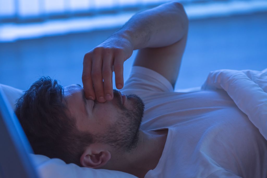 man lying on bed, holding hand on bridge of his nose as he is trying to sleep