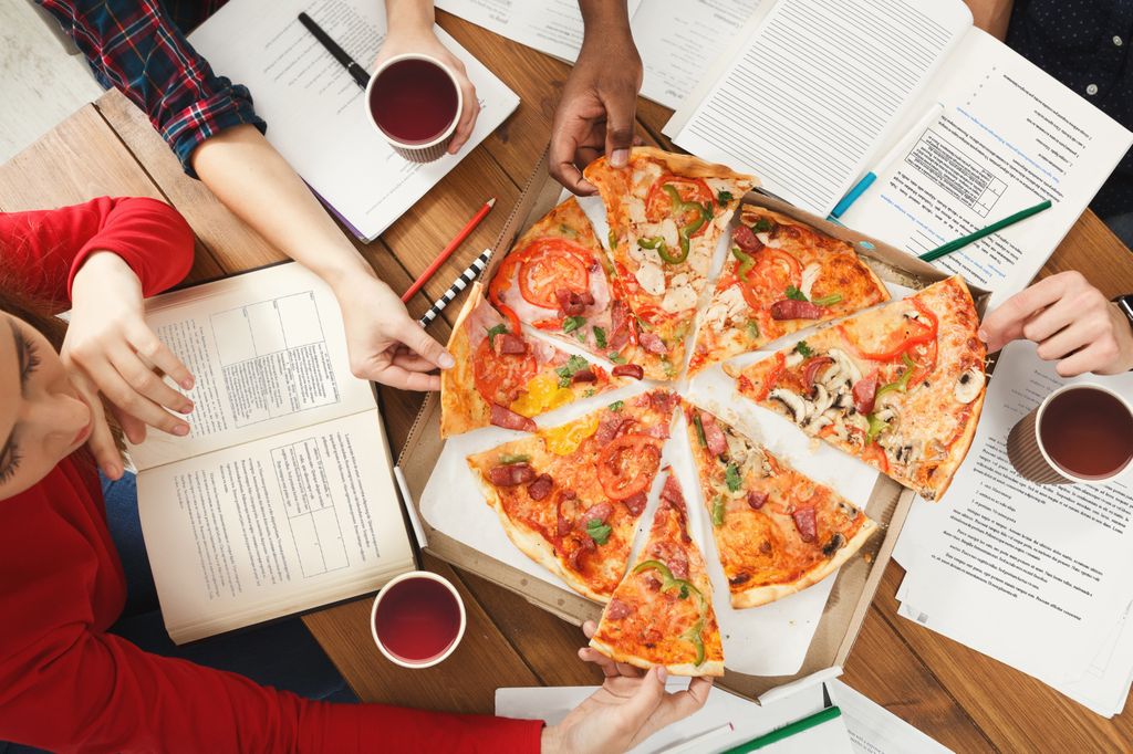 overhead view people sitting around a pizza, with books open, reading, and hands grabbing slices of pizza