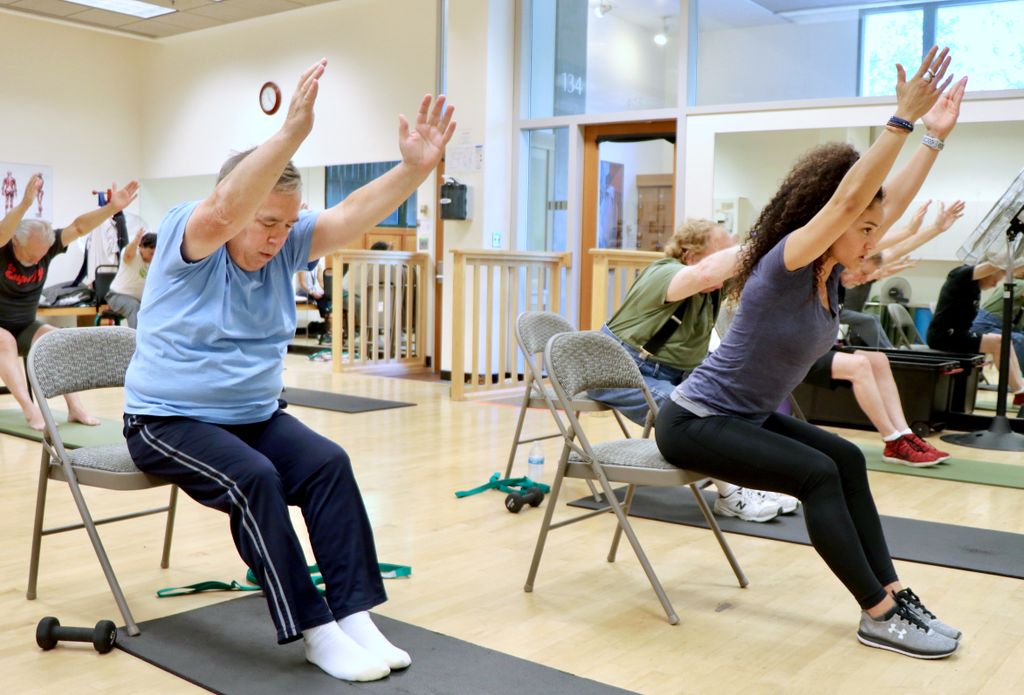 Man, seated to the left, follows a trainer, seated in a chair on the right, leaning forward with arms stretched toward ceiling