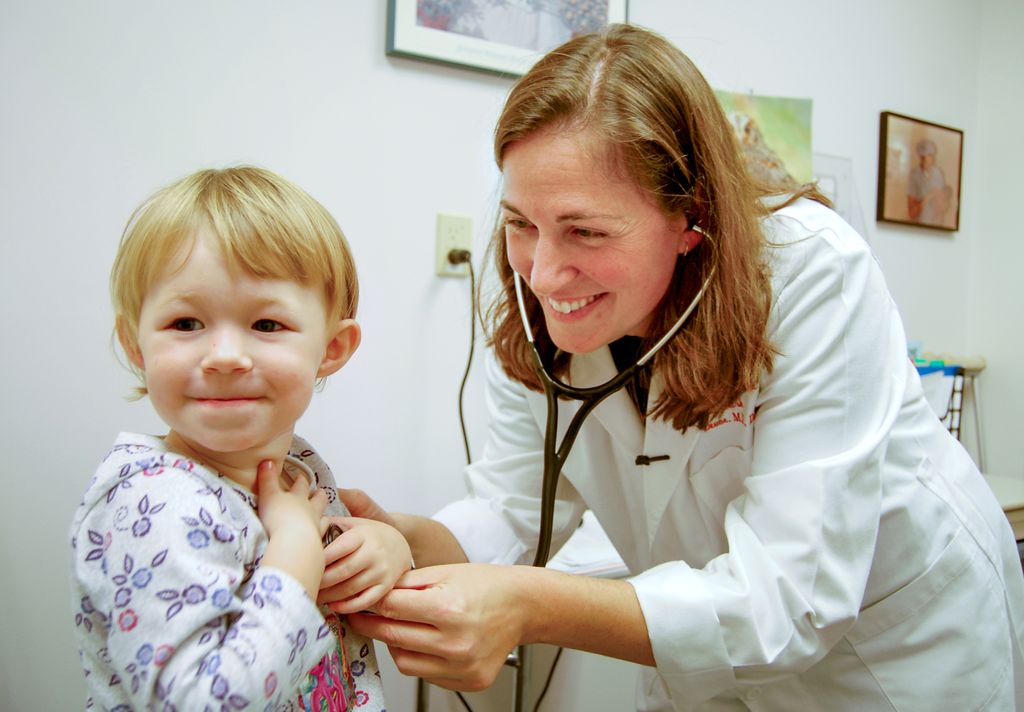 toddler, left, smiling at camera, while doctor, white, with stethoscope around neck, smiles as she tries to listen to child's heart