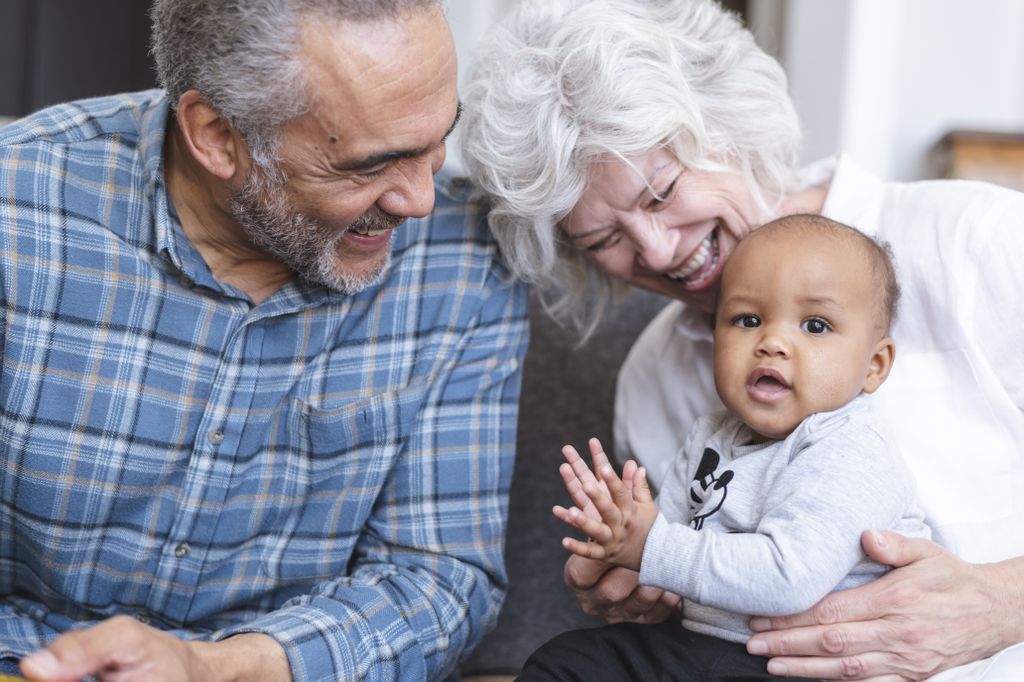A retired senior couple is babysitting their grandchild. The couple is laughing and holding the baby while sitting on the couch. The playful child is clapping.