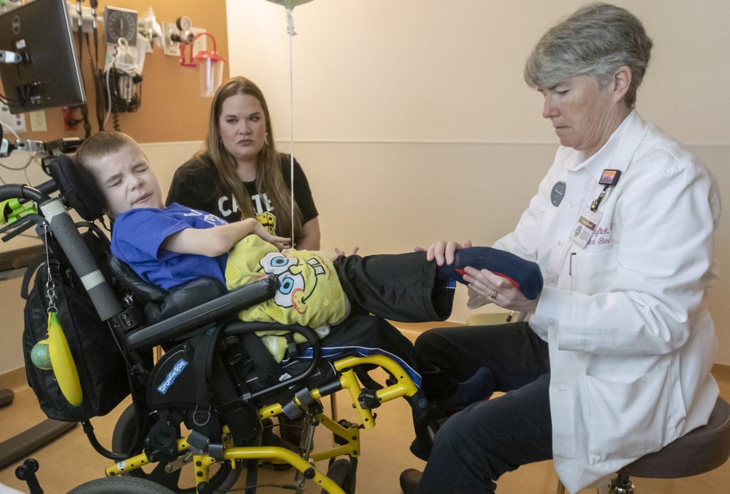 Carter sits in his wheelchair as doctor hayflick, wearing a white coat, seated, to the right, examines his legs