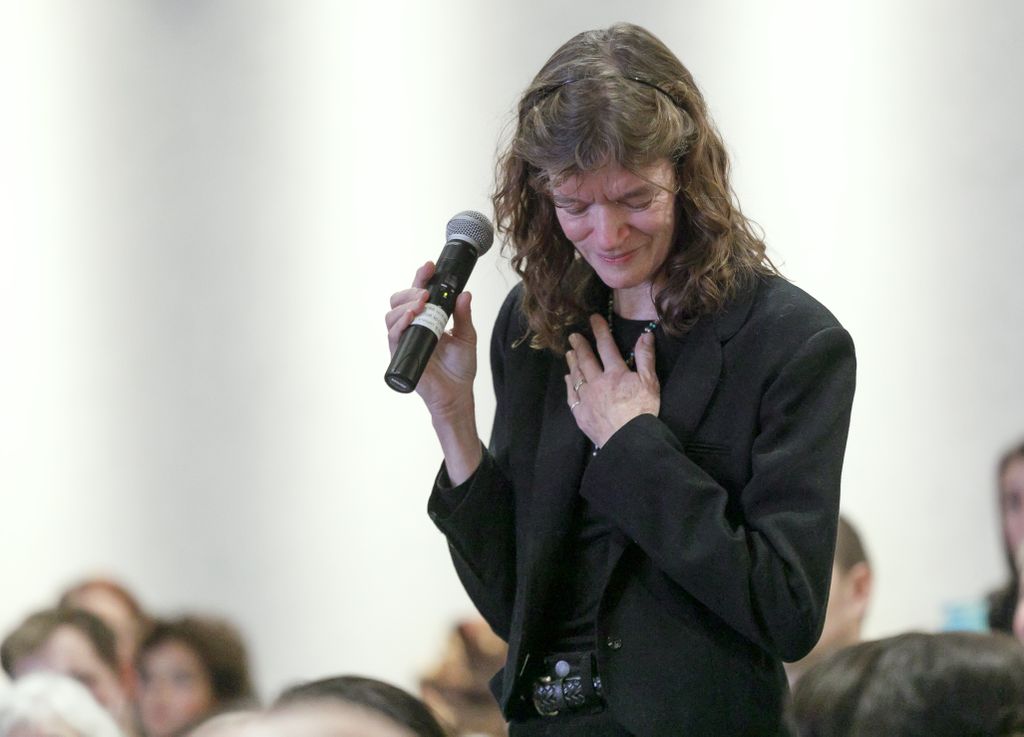 woman holding a microphone, head down, hand on chest over heart