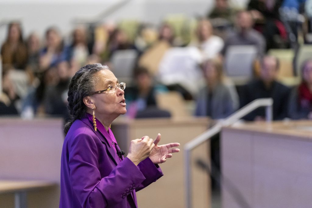 Jones, wearing a purple suit, standing before a large crowd of people as she lectures