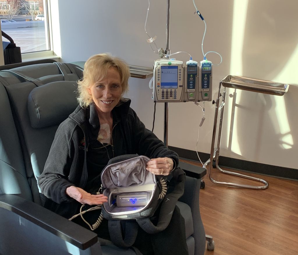 A thin blonde woman in a black jacket sits in a recliner with a machine in the backpack on her lap in a medical clinic