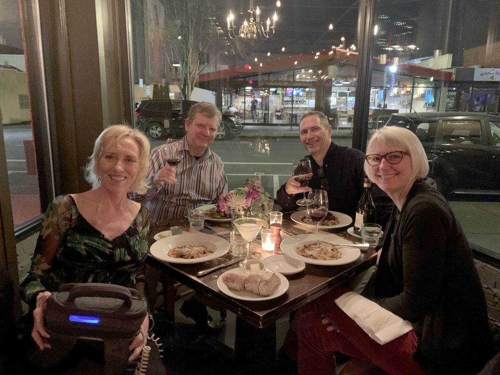 Two men and two women sit around a candlelit dinner table; the blonde woman in the foreground has a glowing machine in a backpack