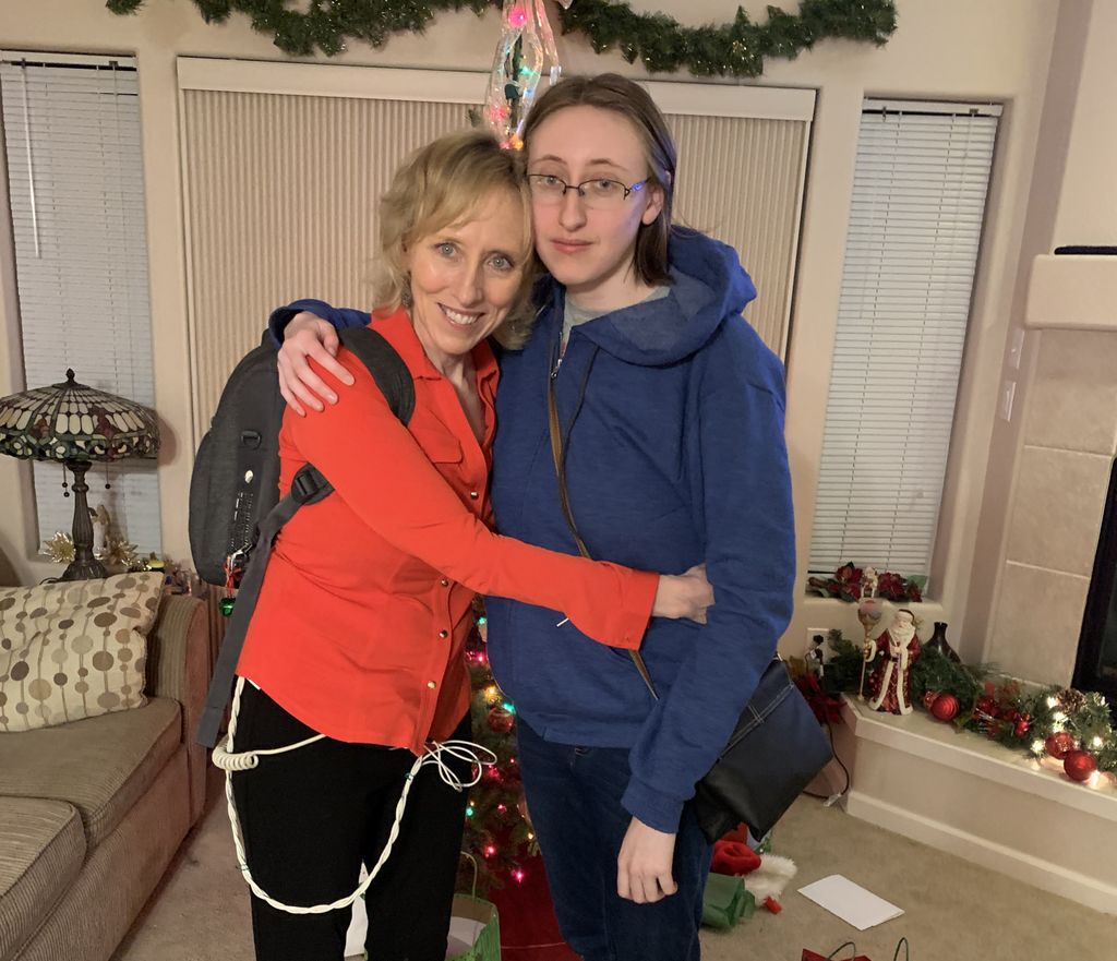 A blonde woman in a red sweater embraces her adult daughter, who has glasses and a ponytail