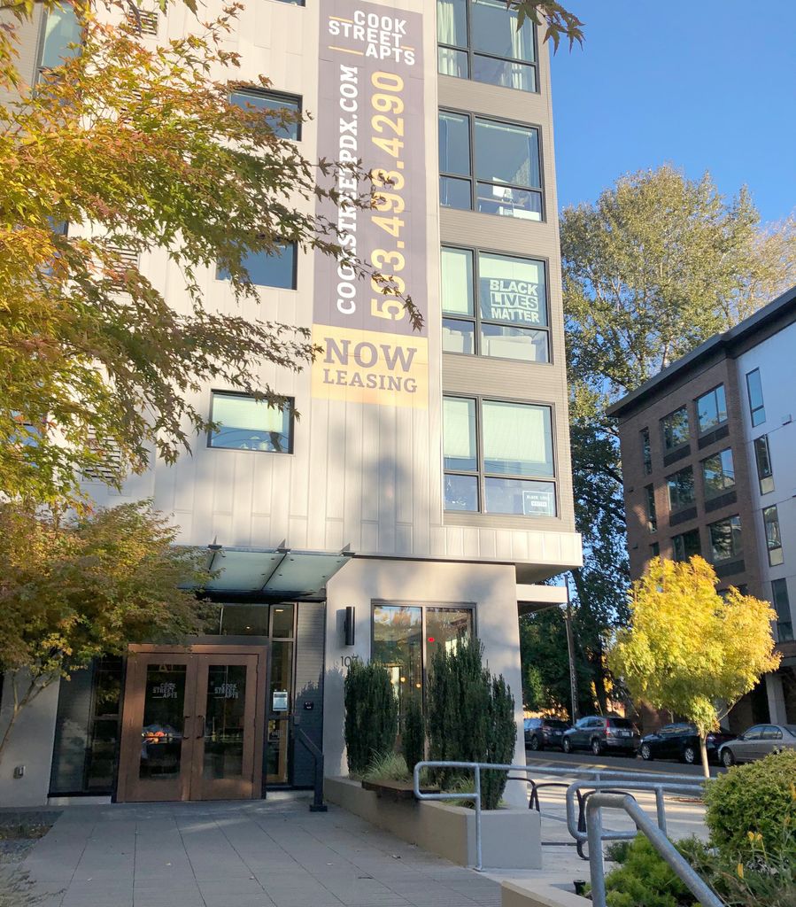 Outdoor, street-level shot of the Cook Street Apartments, a tall, beige building with a 'now leasing' sign in Portland, OR.