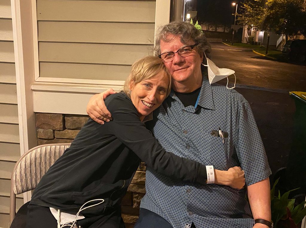 A thin blonde woman has her arms around a grey-haired, smiling man as they sit side by side