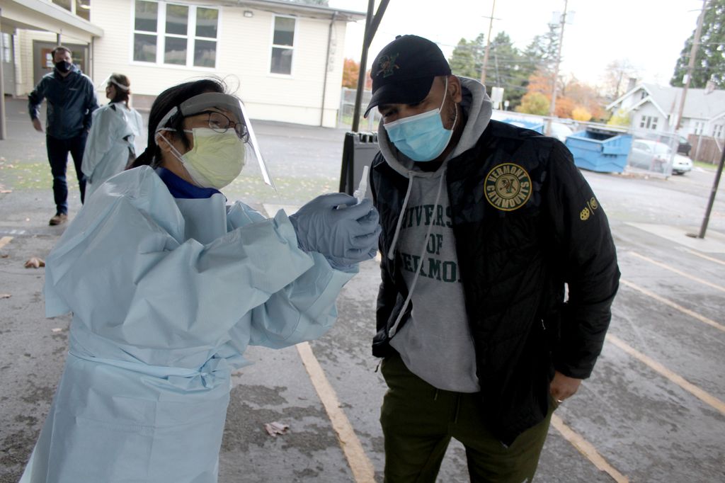 Asian man in full Personal Protective Equipment and masked Black man look at a saliva collection vial