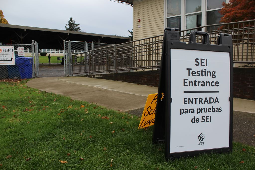 A sign reading "SEI Testing Entrance | ENTRADA para pruebas de SEI" sits on the lawn in front of a building with a ramp entrance