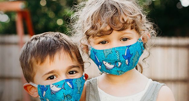 Two young children, a brown-haired boy and blonde girl, stand next to each other while wearing protective masks.