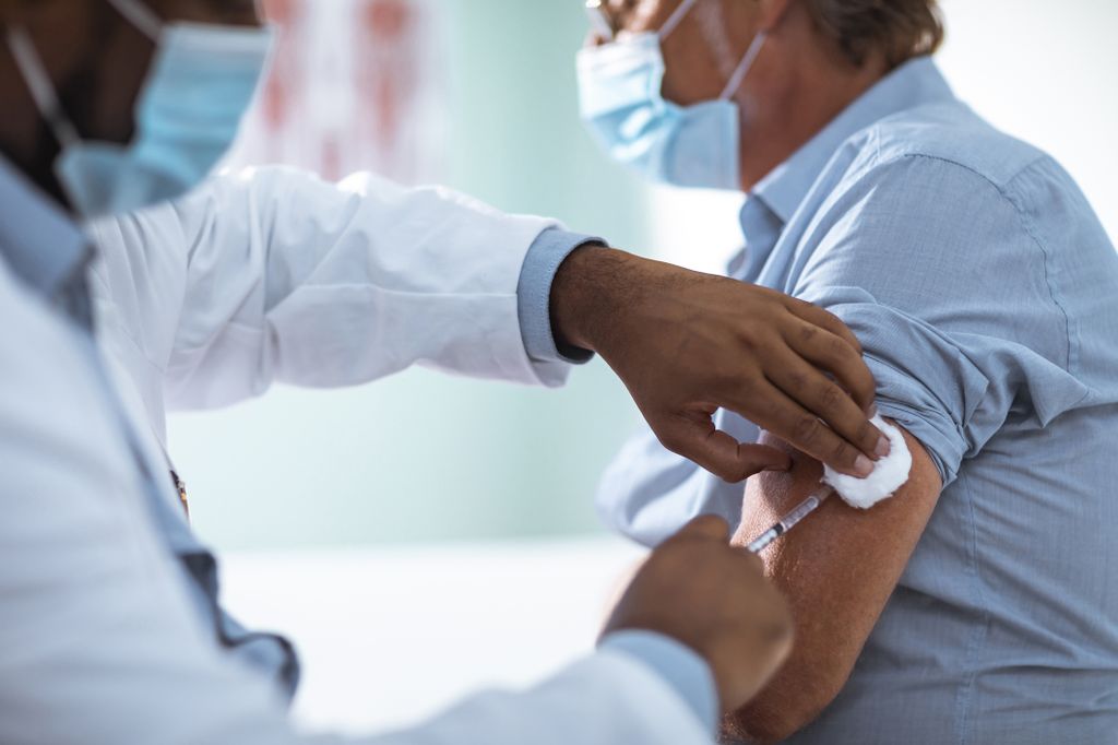 a masked man receives an injection from his masked Black male doctor into his upper left arm