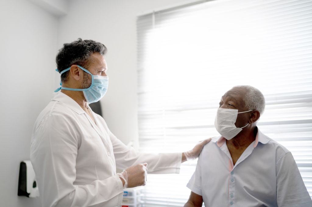 A masked, standing male health care professional in a white lab coat puts his hand on the shoulder of a seated Black male patient