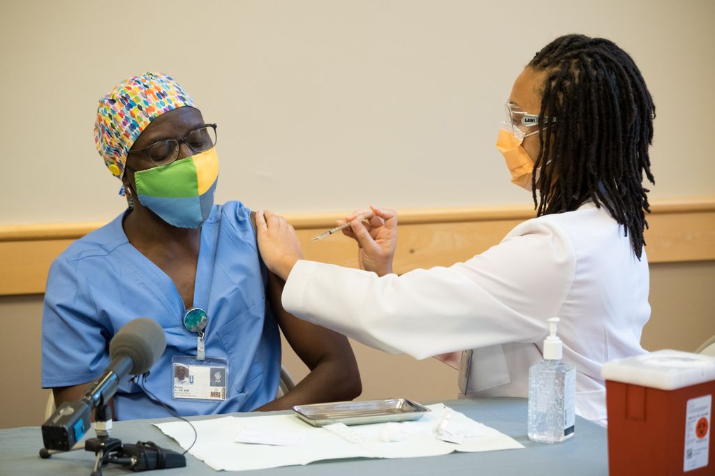 A masked Black adult in scrubs receives an injection from another adult with dreadlocks wearing scrubs 