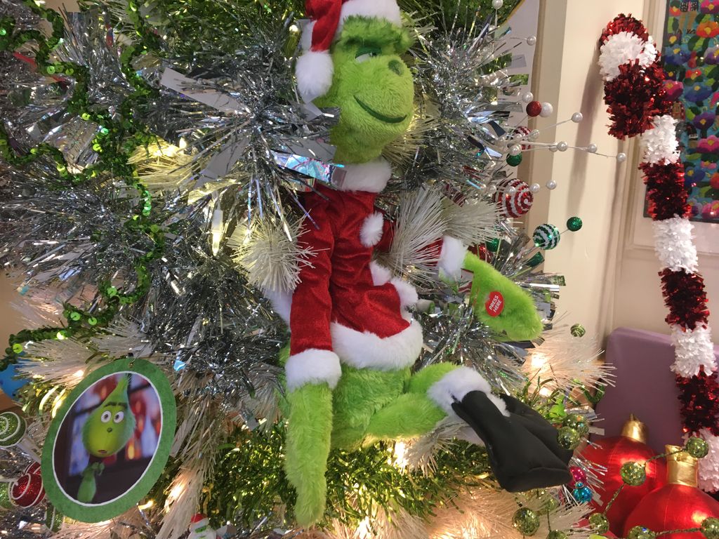 A large plush Grinch (stuffed Dr. Seuss character) and colored lights decorate a Christmas tree; an oversized decorative candy cane leans against the wall